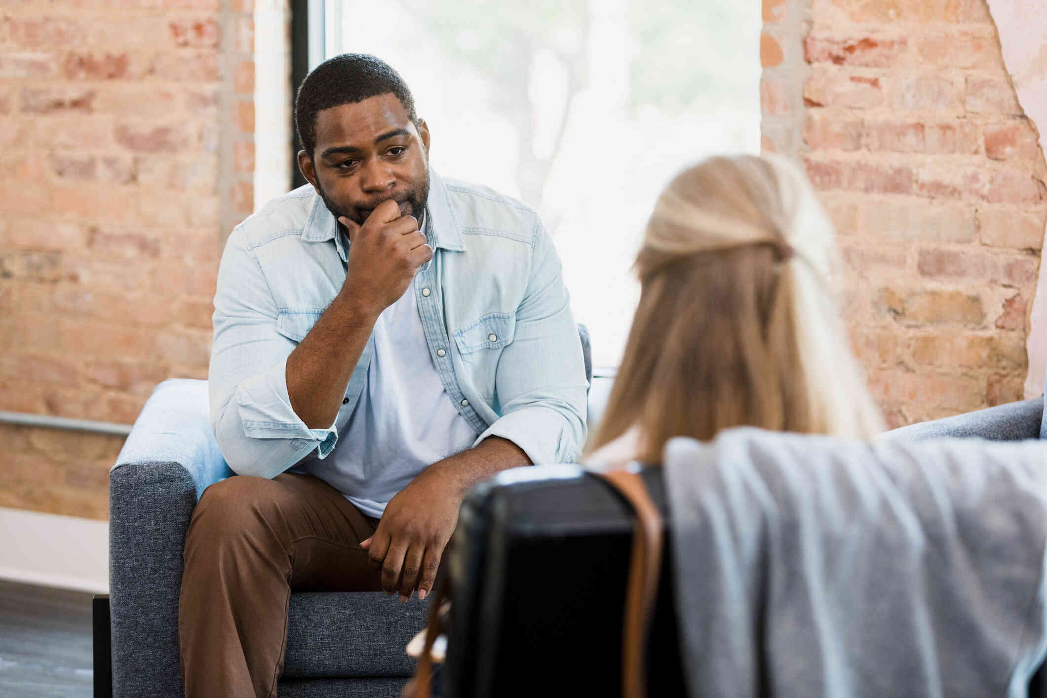 A man in a blue button down shirt sits hunched over in a chair with his hand pressed his face with concern as he listens to the therapist sitting across from him.